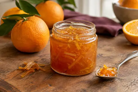 Pot ouvert de marmelade d’oranges amères avec écorces, servi sur une table en bois près d’une tranche de pain.