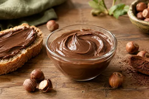Pâte à tartiner chocolat-noisette onctueuse dans un bol, avec une tartine, des noisettes et du cacao
