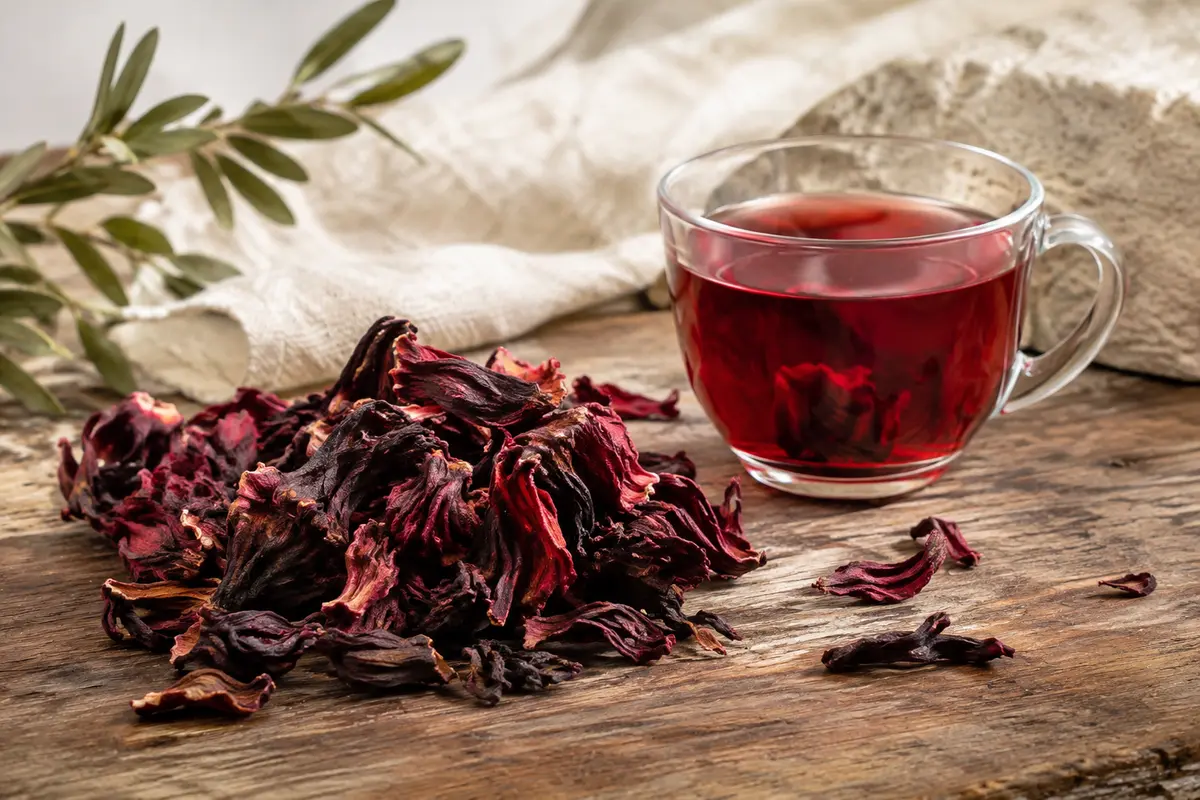 Pétales séchés d’hibiscus dans un bol, près d’une tasse d’infusion rouge sur une table en bois