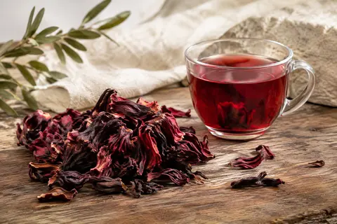 Pétales séchés d’hibiscus dans un bol, près d’une tasse d’infusion rouge sur une table en bois